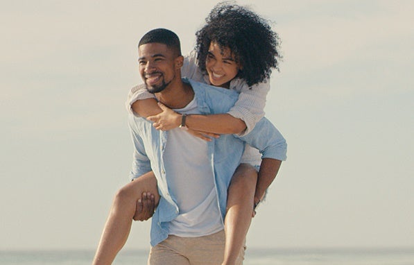 Un couple sur la plage qui regarde au loin, l'homme portant la femme sur son dos.